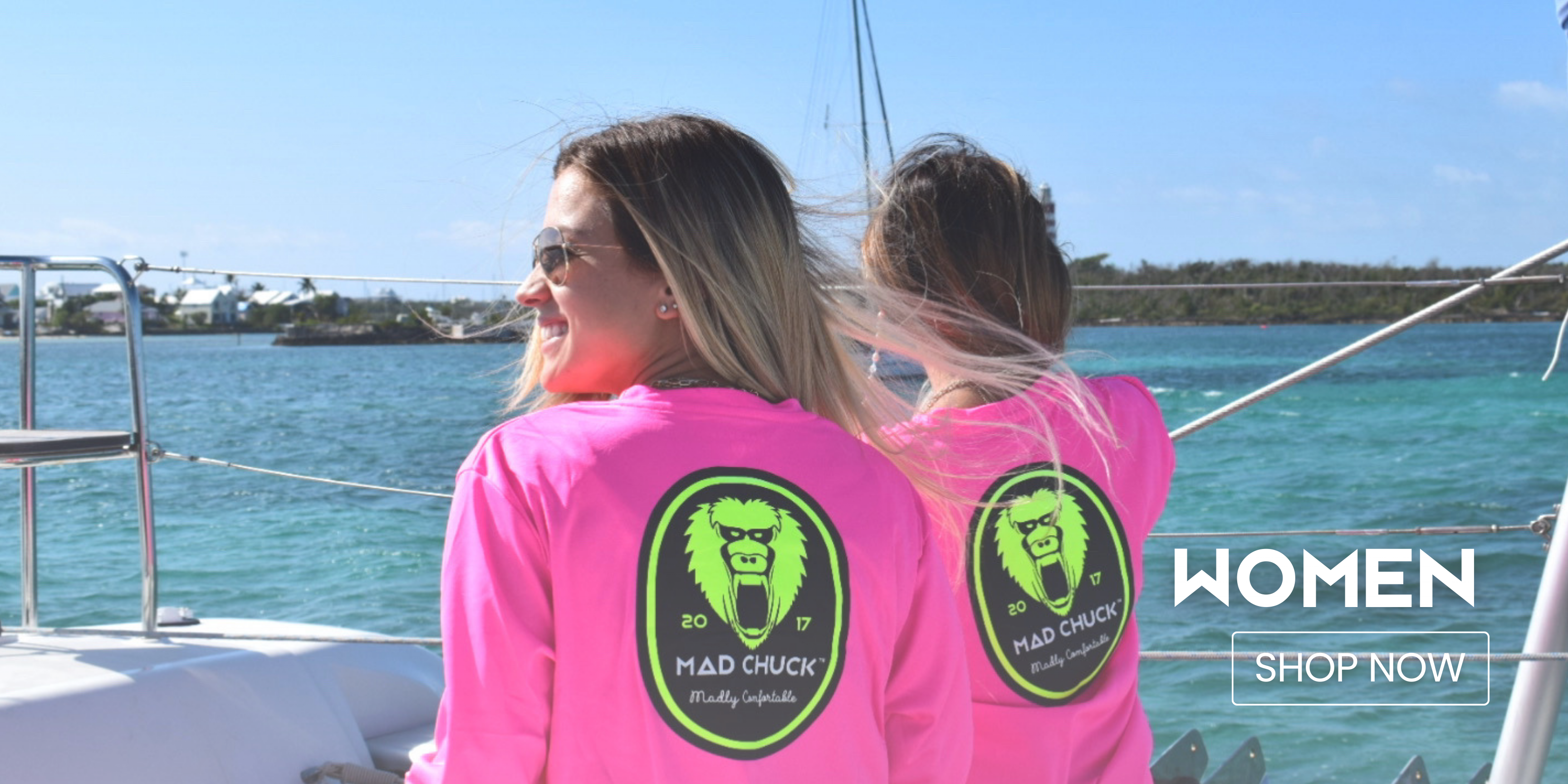 Women - Shop Now: Woman wearing a bright pink Mad Chuck Rashguard sitting on a boat, smiling, with water and a sailboat in the background. Click to browse Women Collection.